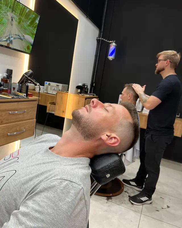 Cliente relaxando na cadeira reclinada enquanto o barbeiro atende ao fundo