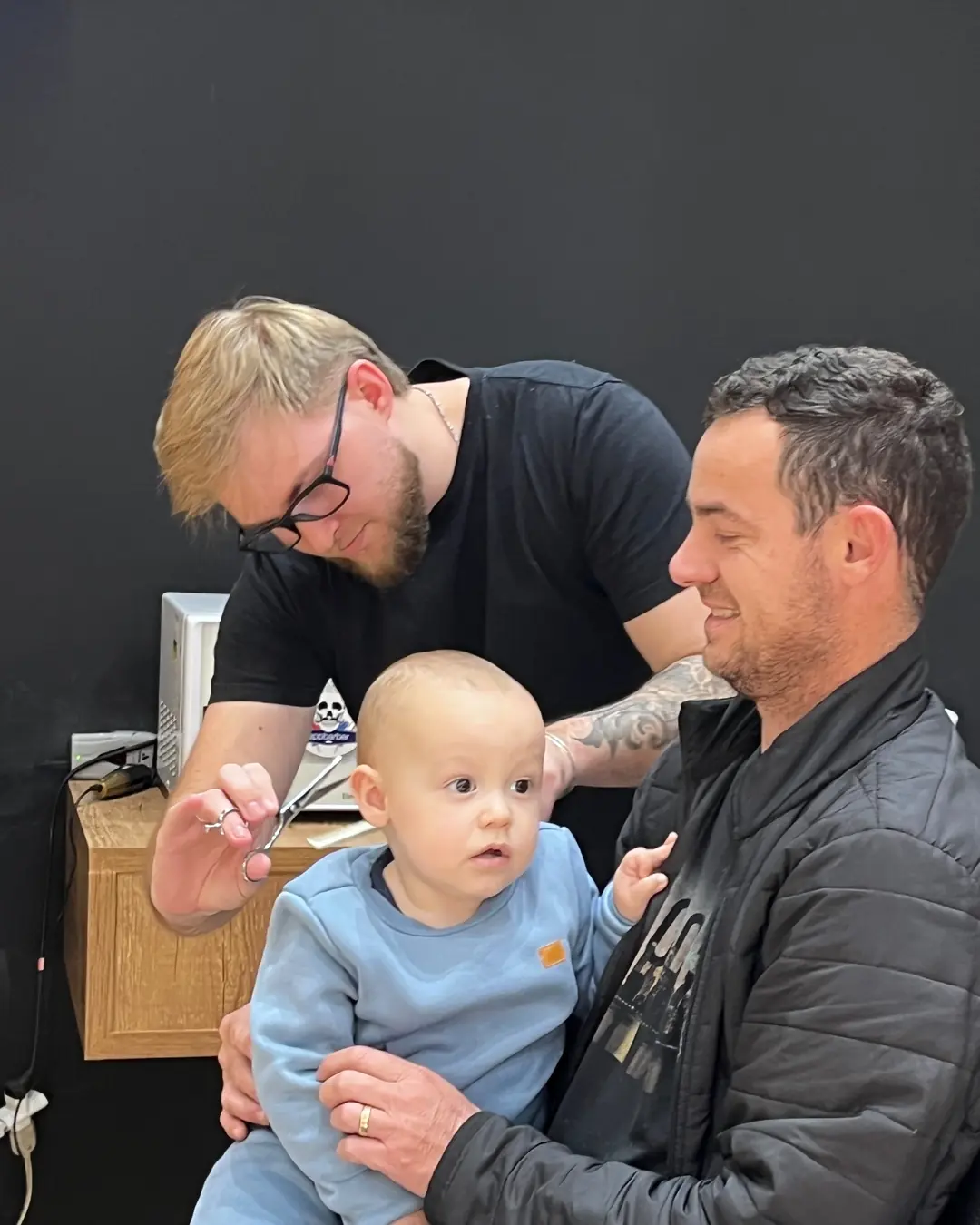 Bebê no colo do pai durante o corte de cabelo, com o barbeiro usando a tesoura